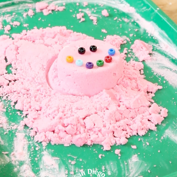 A pile of pink kinetic sand with beads making a smiley face.